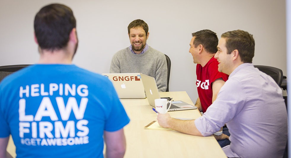 GNGF law firm marketing employees laughing while working around a table. The person in front has a shirt that reads "Helping Law Firms #GetAwesome".